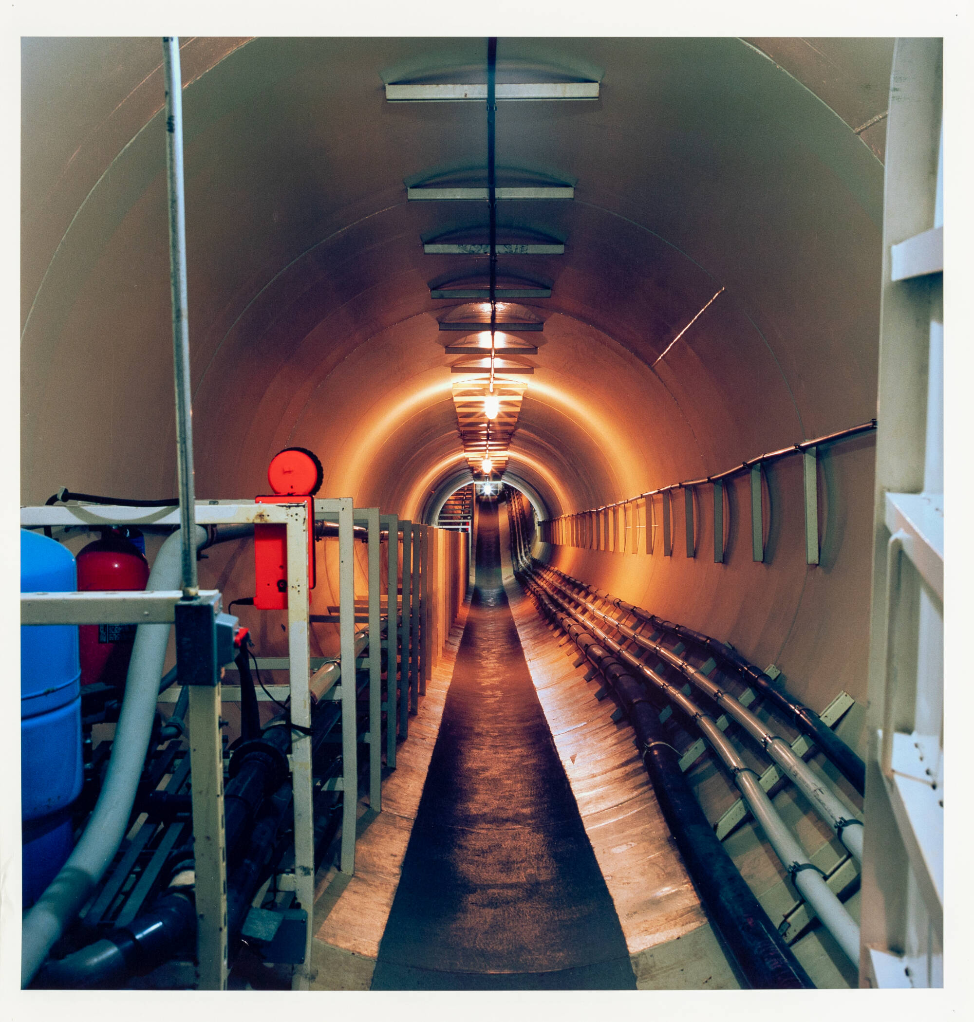 Photograph of the inside of an industrial tunnel with orange lights towards the end of the tunnel.